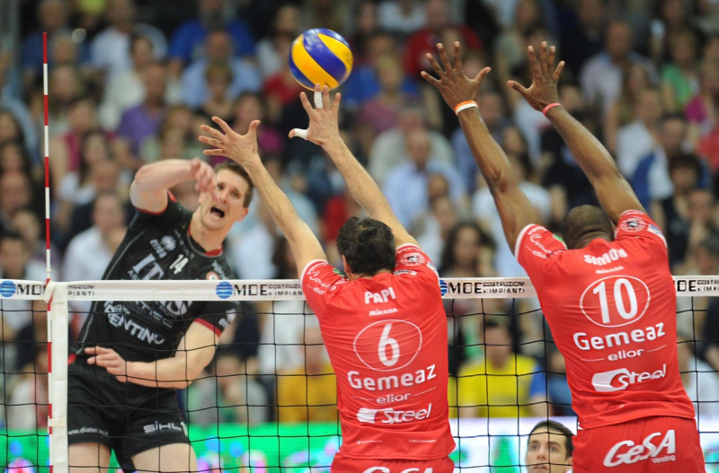 A volleyball player in a black uniform attempts a spike while two players in red jerseys jump to block, near the net during a match.