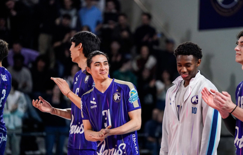Volleyball players in purple jerseys and one in a white jacket stand together smiling and clapping, with a blurred audience in the background.