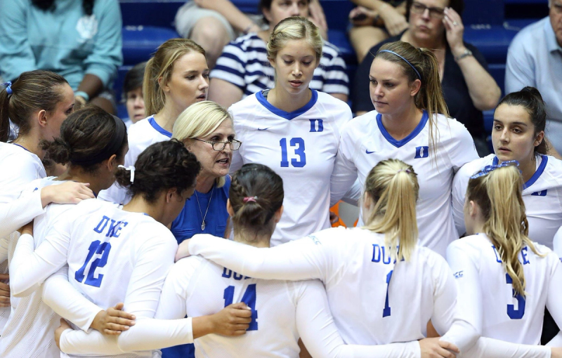A women's volleyball team huddles together with their coach, who is speaking to them. The players are wearing white uniforms with blue accents, and they are listening intently.