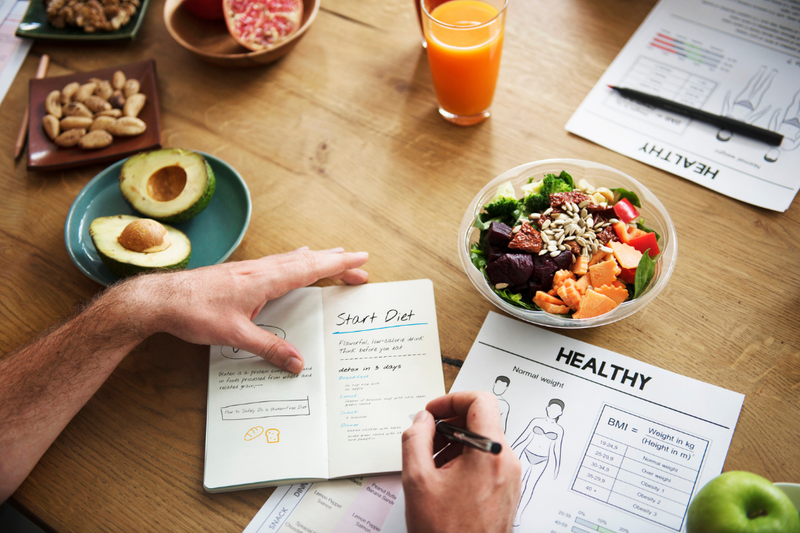 A person writing in a diet journal at a table with healthy food items, including a salad bowl, avocado, almonds, and an orange drink. There are nutrition and health information papers nearby.