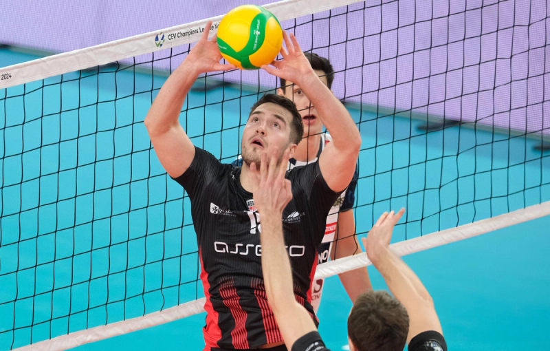 A volleyball player in a black and red uniform is setting the ball near the net during a match. Teammates are visible around, focused on the play.