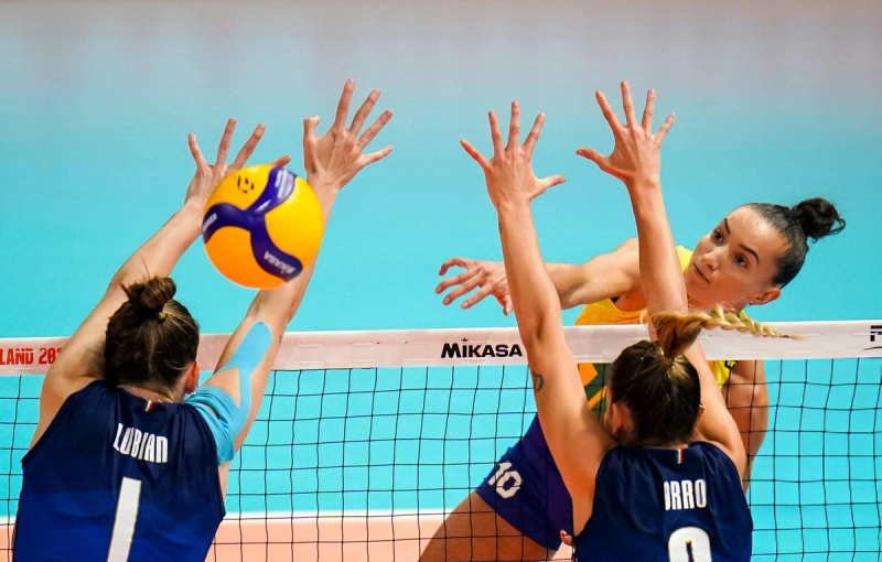 A volleyball player in yellow spikes the ball over the net against two opposing players in blue jerseys who are attempting to block.