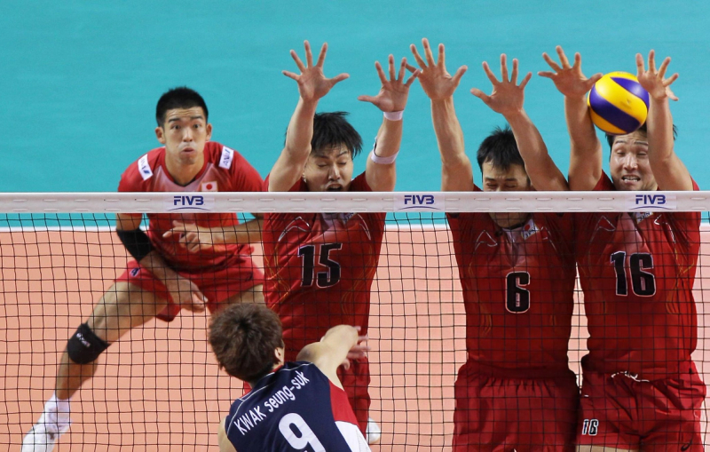 Volleyball match action with three players in red jumping to block a spike from an opposing player wearing a blue jersey.