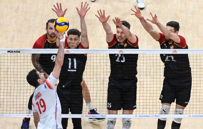 A volleyball player in a white jersey prepares to spike the ball over the net against a four-man block from the opposing team wearing black jerseys.