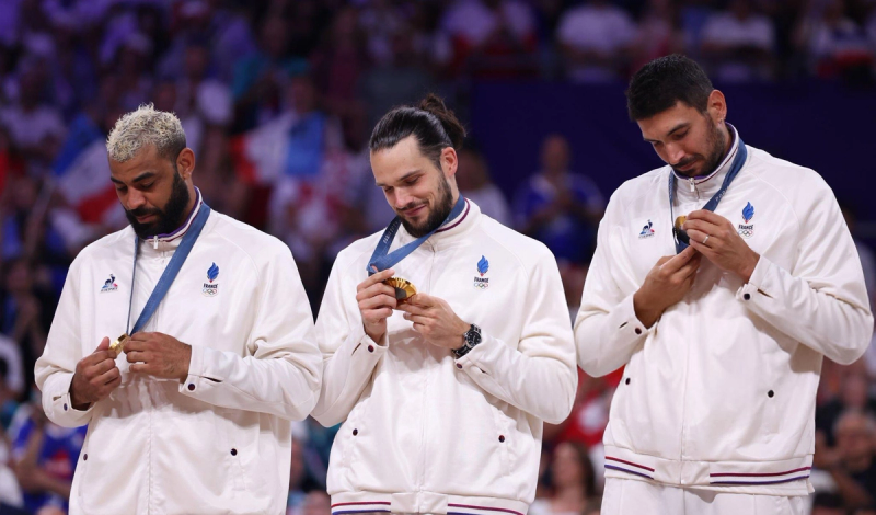 Three athletes from France, wearing white jackets with the French Olympic logo, look at their gold medals during an award ceremony.
