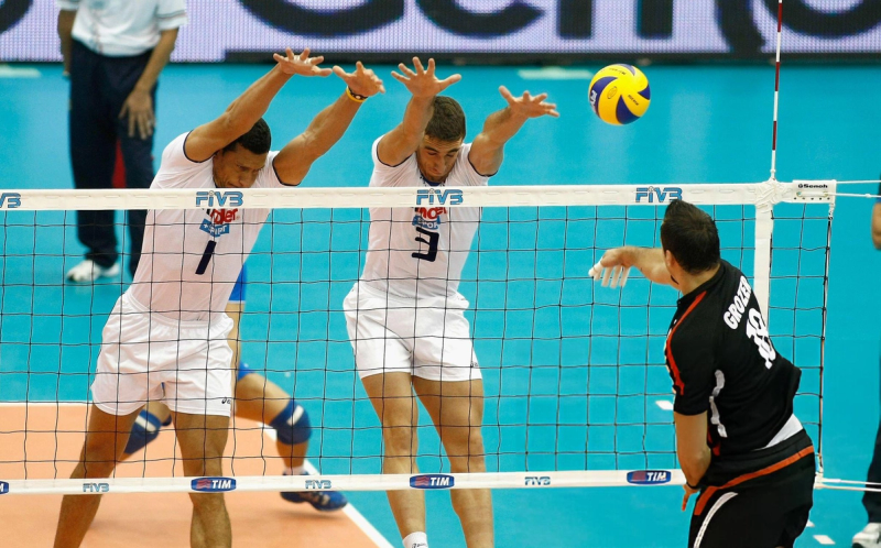 Two volleyball players in white jerseys attempting to block a ball at the net against a player in a black jersey during a match.