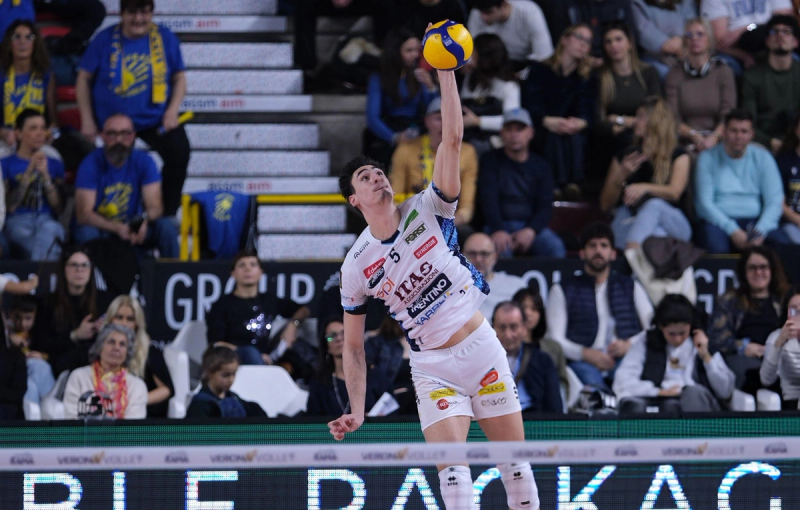 A volleyball player in a white uniform is leaping into the air to hit a ball. The crowd is visible in the background, watching the game intently.