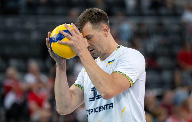 A volleyball player in a white jersey holds a volleyball against his forehead, appearing focused and contemplative during a match.