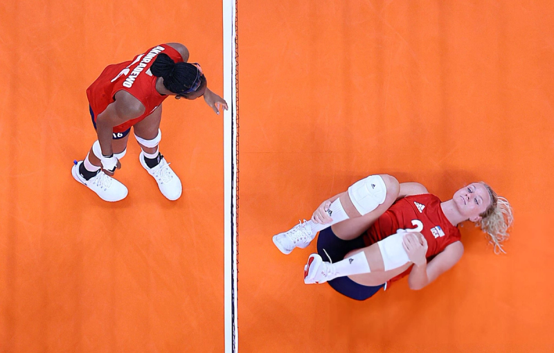 Two volleyball players on an orange court; one is standing and looking at the other, who is lying on the ground holding their knee.