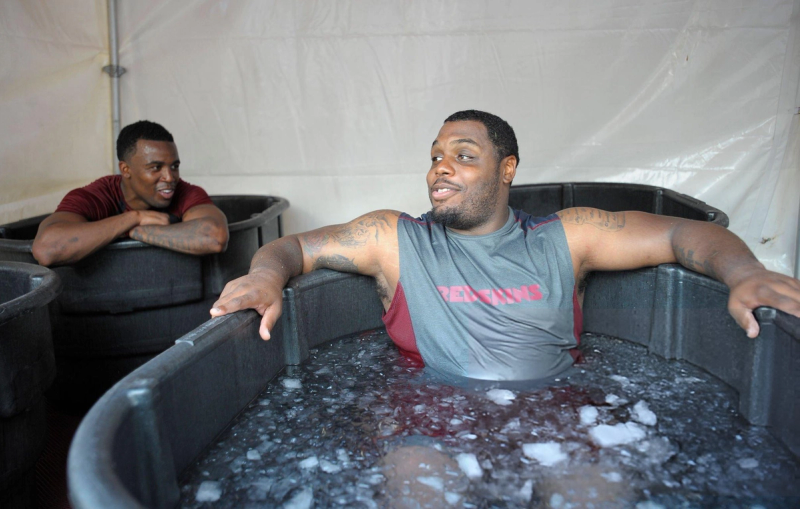 Two men are smiling and talking while sitting in separate ice baths. They are inside a tent, with one man showing visible tattoos.