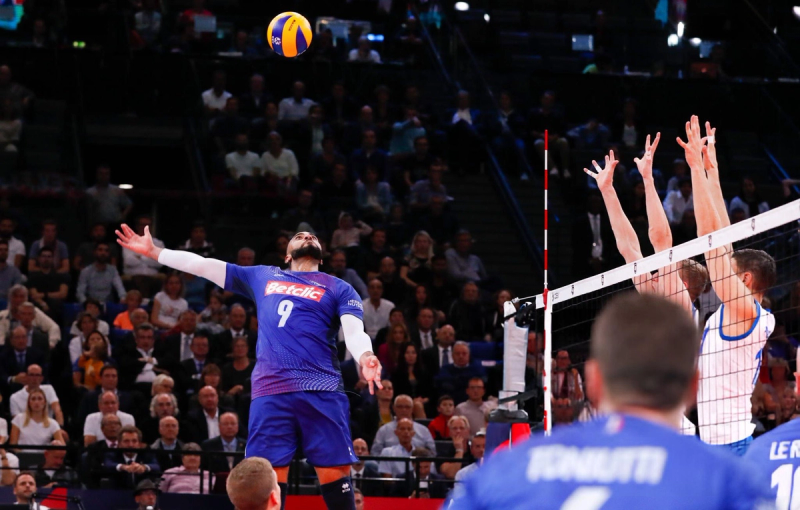 Volleyball player in a blue jersey prepares to spike the ball as two opponents in white jerseys attempt to block across the net. Spectators are visible in the background.