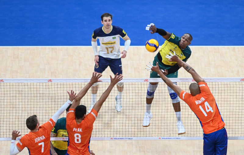 A volleyball player in a yellow and green uniform spikes the ball over the net as three players in orange jerseys attempt to block. Another player in a blue and white uniform watches from the back.