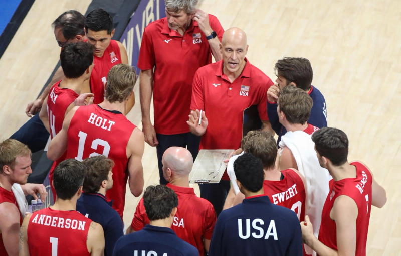 A volleyball coach giving instructions to his team, who are wearing red jerseys with "USA" on them, during a timeout. The players are gathered closely, listening attentively.