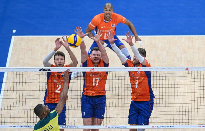 Volleyball match showing three players in orange jerseys attempting to block a spike from an opposing player in a yellow jersey at the net.