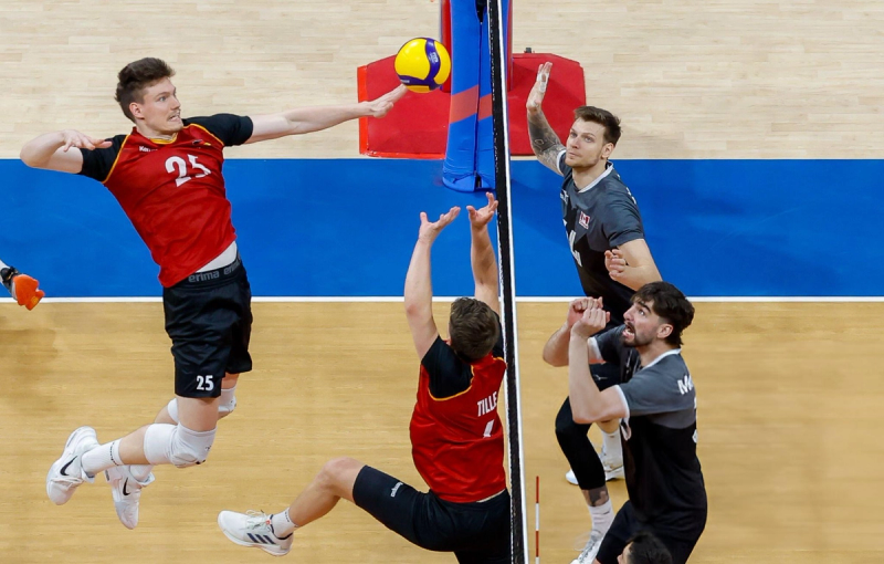 A volleyball player in a red jersey jumps and prepares to spike the ball over the net, while opponents in black jerseys attempt to block. The court is wooden with blue markings.