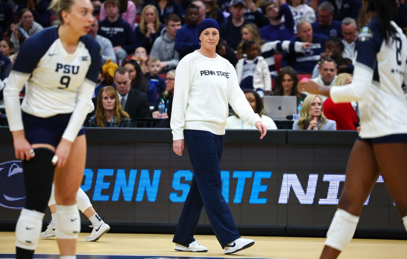 A person wearing a "PENN STATE" sweatshirt and blue pants is standing on a volleyball court. They are surrounded by players in PSU volleyball uniforms, and there is a crowd sitting in the background.