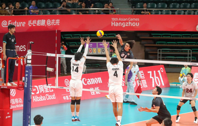 Volleyball match at the 19th Asian Games in Hangzhou. A player in a dark jersey spikes the ball over the net against two opposing team players in white jerseys attempting to block. An official observes the action, with a red event banner in the background.