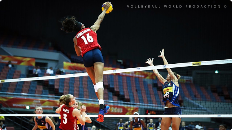 A volleyball player in a red jersey leaps high above the net, reaching to hit the ball, while players in blue attempt to block. Stadium seats are visible in the background.