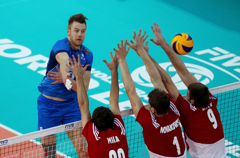 A volleyball player in a blue jersey prepares to spike the ball over the net, while three players in red jerseys attempt to block. The court's surface is turquoise, and the FIVB logo is visible.