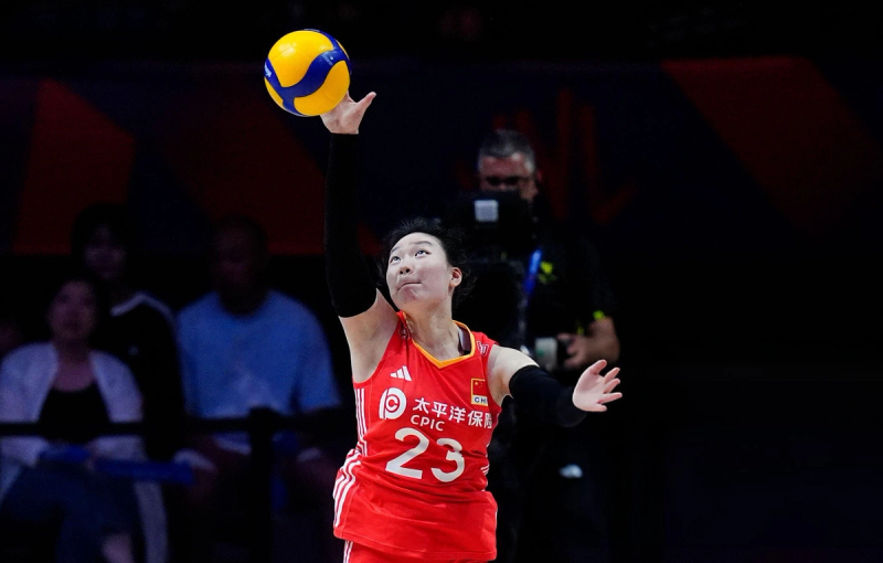 A volleyball player in a red jersey, number 23, is about to serve the ball during a match. The background shows some spectators and a cameraman.