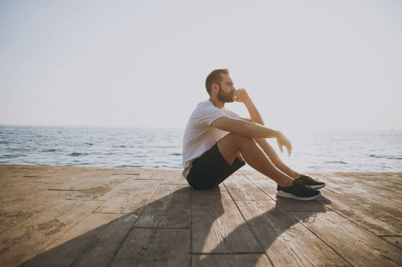 A person sitting on a wooden dock by the sea, gazing thoughtfully at the horizon, wearing a white t-shirt, black shorts, and sneakers, in bright sunlight.