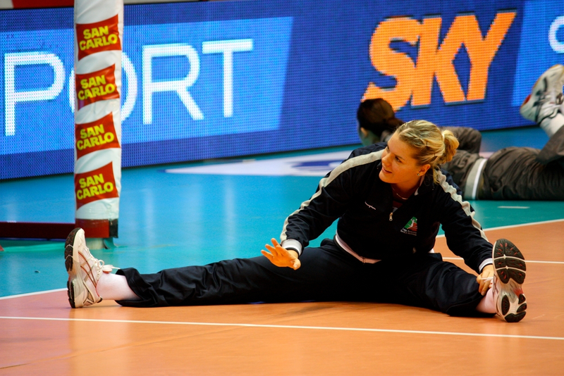 A volleyball player stretches on the court, sitting with legs apart. She wears a tracksuit and is smiling, preparing for a match. The court is marked with brand logos in the background.