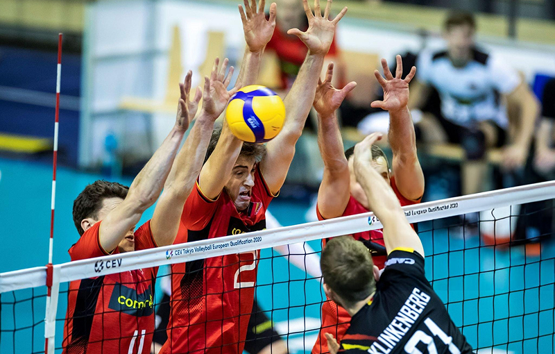 Players in red jerseys attempt to block a volleyball at the net, while a player in a black jersey jumps to hit it during a competitive match.