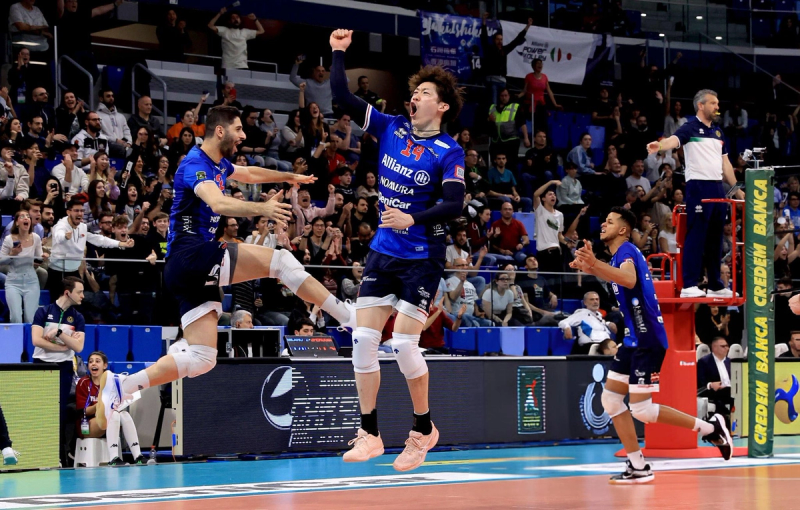 Volleyball players in blue jerseys celebrate a point on the court, with a cheering crowd in the background.