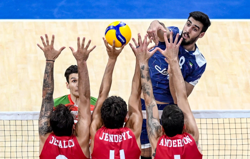 A volleyball player in a blue uniform spikes the ball over the net against a team in red jerseys, with three opposing players attempting to block the shot.