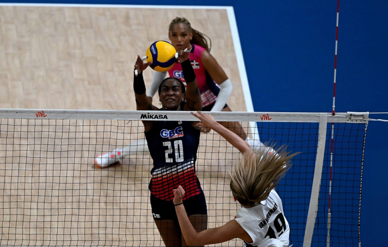 Two volleyball players competing at the net during a match, with one attempting a block and the other spiking the ball. A referee is visible in the background.