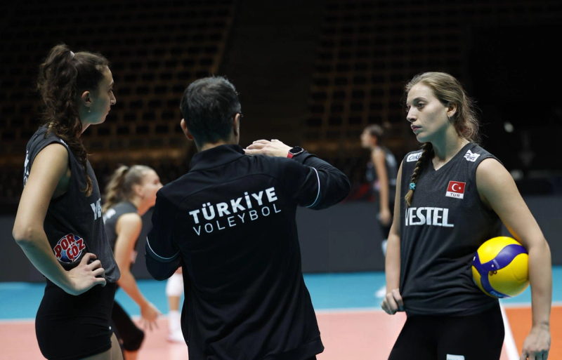Volleyball players from Türkiye's team listen to a coach during a break on an indoor court. One player holds a volleyball, and their jerseys display the Turkish flag.