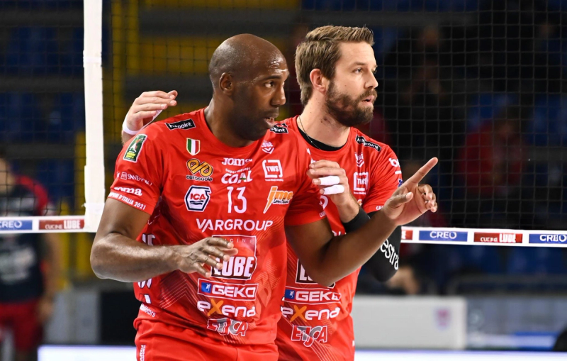 Two volleyball players in red jerseys on the court, focused, with one pointing and the other gesturing supportively.