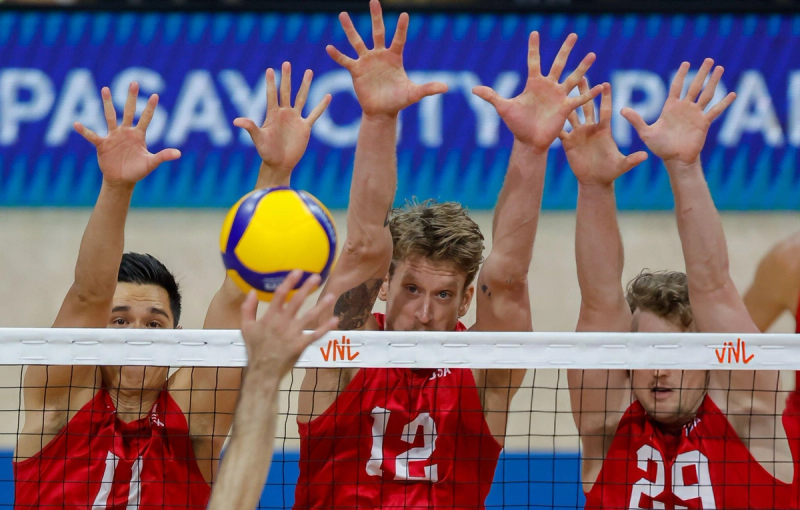 Three volleyball players in red uniforms jump to block a ball at the net during a match.