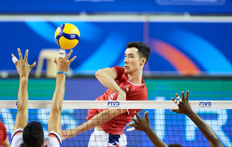 Volleyball player in a red jersey spiking the ball over the net while opposing players attempt to block, during an indoor match.
