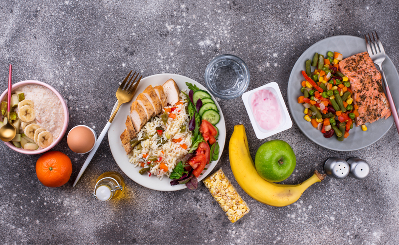 A selection of foods including oatmeal with banana slices, a tangerine, boiled egg, chicken with rice and salad, salmon with mixed vegetables, yogurt, an apple, banana, granola bar, and a glass of water on a textured surface.
