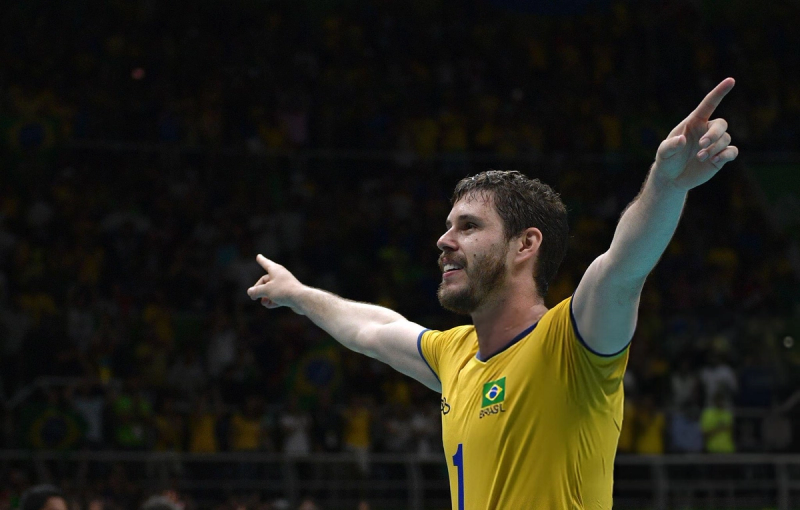 A volleyball player in a yellow jersey with the Brazilian flag is celebrating with arms outstretched, pointing upwards, in a sports arena with a crowd in the background.