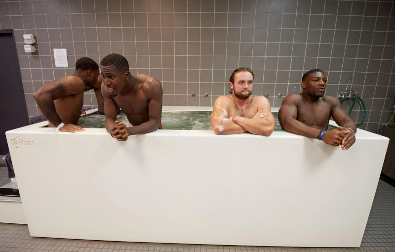 Four men are in a rectangular hot tub. One has his back turned, two are leaning on the side, and one is sitting with arms crossed. The room has tiled walls and a floor.