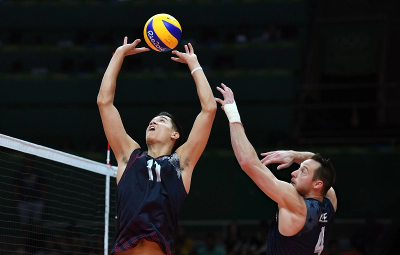 Two volleyball players in dark jerseys are in action. One player is setting the ball while the other prepares to spike it. The ball is marked with "Rio 2016."
