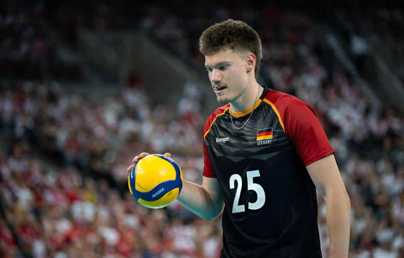 A volleyball player in a black and red Germany jersey, numbered 25, holding a volleyball. The background is a blurred crowd.