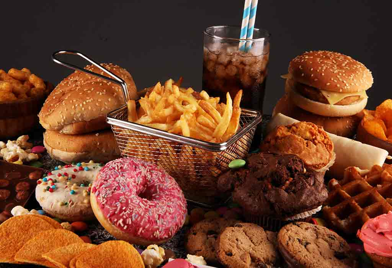 A selection of fast food and desserts, including burgers, fries in a basket, a glass of soda with straws, donuts with sprinkles, cookies, muffins, potato chips, and waffles, all arranged on a dark background.