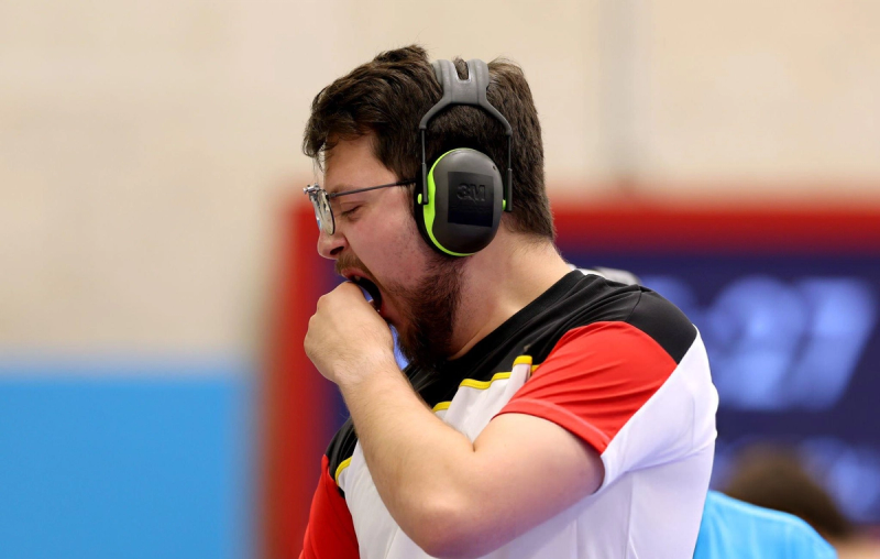 A man wearing earmuffs and glasses, yawning with his hand near his mouth, dressed in a sports jersey.