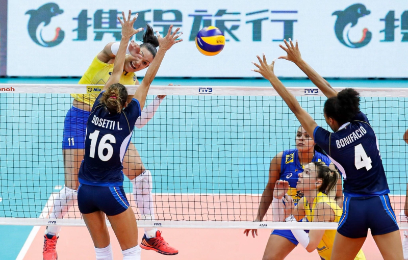 Women's volleyball match with players in action at the net, one in a yellow jersey attempting to spike the ball while two others in blue jerseys attempt to block.