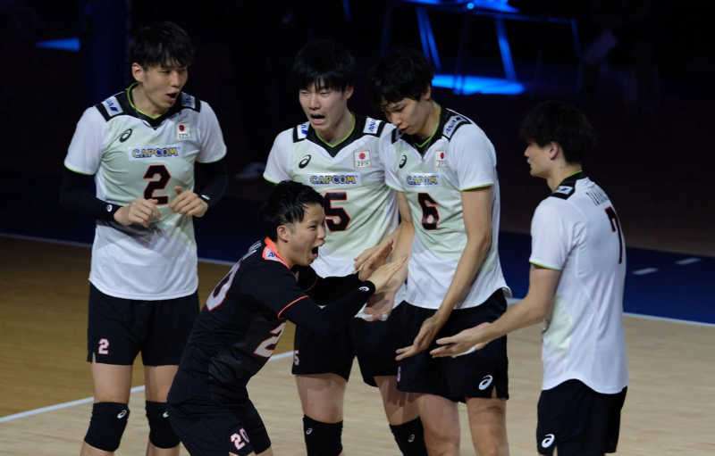 A group of Japanese volleyball players in team jerseys are gathered on the court, showing expressions of excitement and camaraderie.