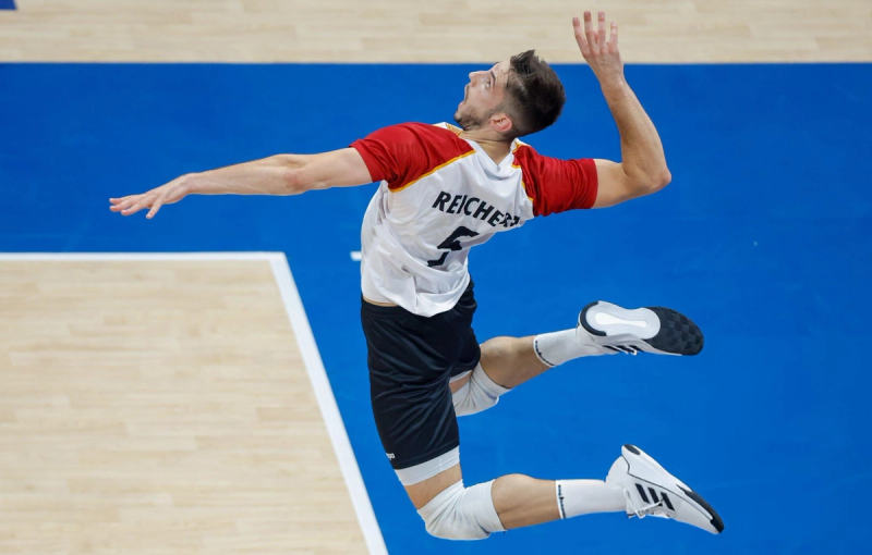 A volleyball player in a red and white jersey leaps mid-air about to spike the ball on a blue indoor court.
