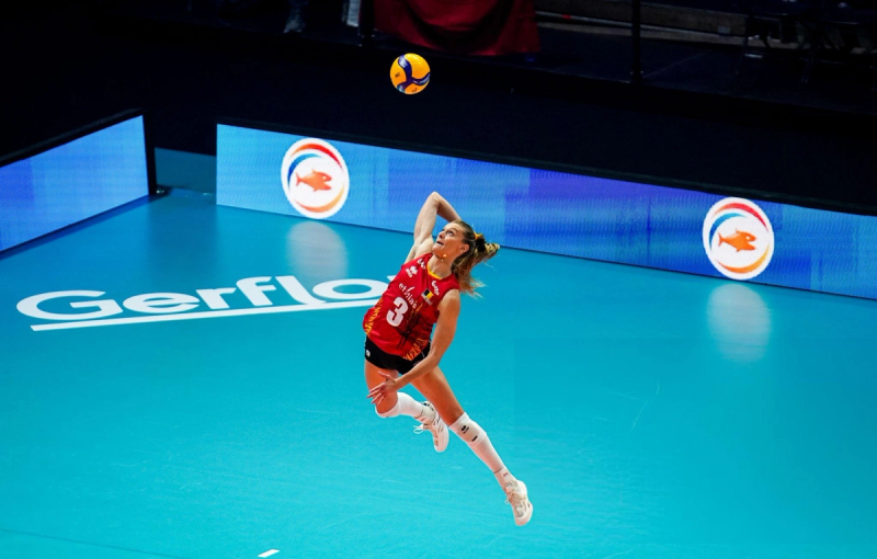A volleyball player in a red jersey leaps into the air to serve the ball on a blue court.