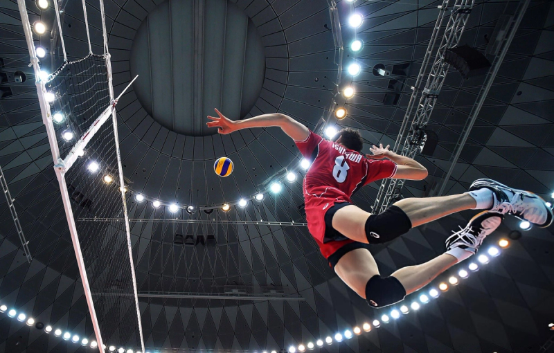 Volleyball player in a red jersey jumps mid-air near the net to hit the ball, with bright arena lights in the background.