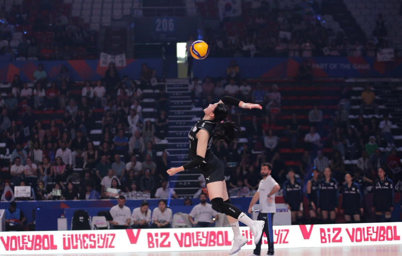 A volleyball player in a black uniform is captured mid-air, preparing to serve the ball. The background shows an arena with spectators watching intently.