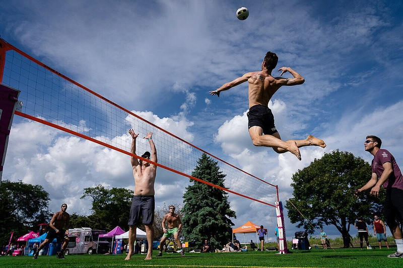 Four men are playing an outdoor volleyball game. One player is airborne, attempting to spike the ball over the net while others are in position. A clear sky and trees are in the background.