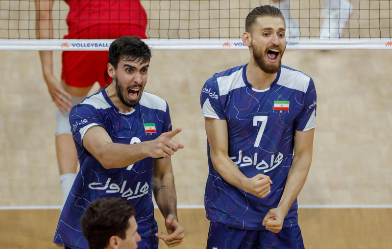 Two volleyball players, wearing blue jerseys with the number 3 and 7, celebrate enthusiastically during a match. A net and players in red are visible in the background.