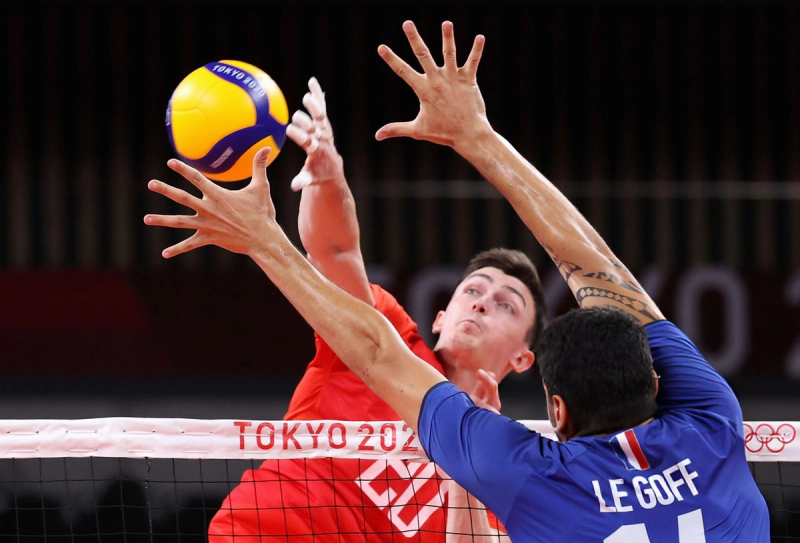 Two volleyball players, one in a red jersey and one in blue, reach for the ball at the net during a match. The venue is identified as "Tokyo 2020."